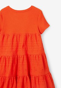 Oranžové šaty s žebrovaným horním dílem a vrstvenou sukní s průstřihy. Látka má texturu s motivy a jemnými detaily po celém povrchu.