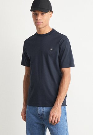 Donkerblauwe t-shirt met korte mouwen van een zachte stof, met een klein logo op de borst en een comfortabele pasvorm, gecombineerd met lichtblauwe jeans.