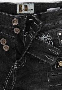 Jeans in denim nero con patta a bottoni, hardware in bronzo, cuciture bianche a contrasto e un design ricamato sulla cintura.