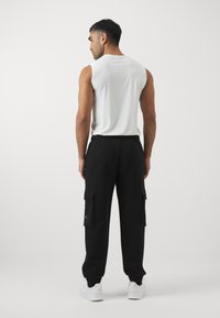 Ein Mann steht mit dem Rücken zur Kamera, trägt ein weißes ärmelloses Shirt, schwarze Cargohosen und weiße Sneakers vor einem einfarbigen Hintergrund.