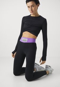 Top cropped preto de mangas longas com um design torcido. Calças leggings pretas com uma faixa roxa na cintura e uma etiqueta de marca. Ténis atléticos cinzentos.