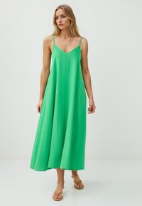 Eine Frau steht in einem langen, ärmellosen grünen Kleid und Sandalen vor einem schlichten hellen Hintergrund. Nachdenklich blickt sie in die Ferne.