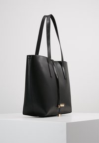 Sac fourre-tout en cuir noir avec deux longues poignées, design minimaliste, forme plate et un accent doré sur le devant pour la fermeture.