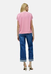 Mujer vestida con una camiseta rosa de manga corta, jeans azules arremangados y sandalias de tacón beige, de pie de espaldas sobre un fondo blanco.