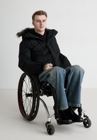 Chaqueta acolchada negra con capucha forrada de pelo, llevada sobre una camiseta negra. Jeans de mezclilla azules holgados, combinados con botas negras, sentado en una silla de ruedas.