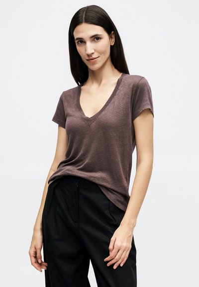 Jeune femme aux longs cheveux foncés portant un t-shirt marron à manches courtes en V et un pantalon noir, sur un fond uni.