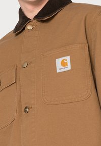 Hnědá plátěná bunda s kordovou límcem, která má kovové knoflíky a viditelnou bílou náprsní kapsu s oranžovým logem.