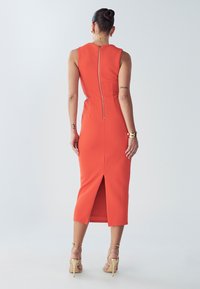 Orange tætsiddende kjole med lynlås bagpå og en midlang kant, der har en slids bagpå. Båret med slidse smalle beige hæle.