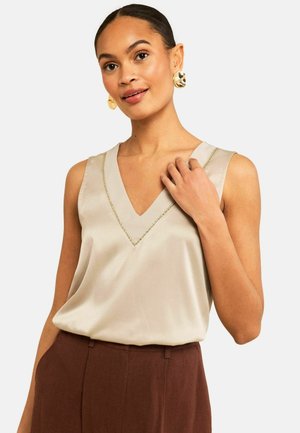 Woman wearing sleeveless beige satin top with V-neck and brown high-waist pants, accessorized with large gold earrings.