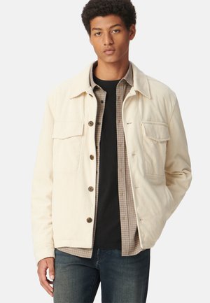 Beige corduroy jas met een kraag, vier zakken en bruine knopen, gelaagd over een geruit shirt, gecombineerd met donkere spijkerbroek.