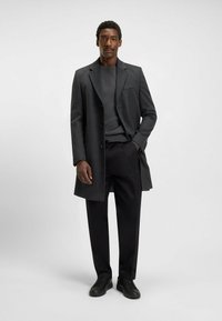 Grijze wolmix-overcoat met een notch-revers, een enkelzijdige sluiting en een borstzak, gedragen met een donker truibroek en zwarte trousers.