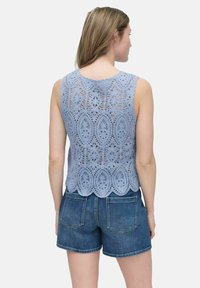 Ljusblå spetstopp med blommönster och scalloped nederkant, bärs över denimshorts. Tyget verkar texturerat och skört.