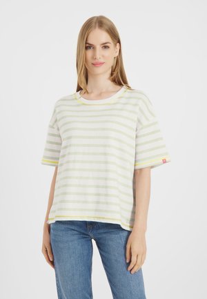 Blondynka ubrana w luźny biały tshirt z jasnozielonymi poziomymi paskami oraz niebieskie dżinsy, stojąca na białym tle.