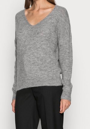 Kobieta ubrana w miękki szary sweter z dekoltem w serek i długimi rękawami, w połączeniu z luźnymi czarnymi spodniami, stojąca na jednolitym tle.