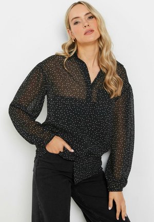 Schwarze durchscheinende Bluse mit weißen Punkten, Knopfverschluss vorne und gerafften Manschetten. Kombiniert mit schwarzen Hosen.