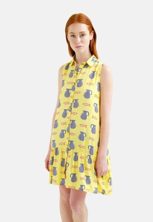 Mujer pelirroja que lleva un vestido amarillo sin mangas con motivos de jarras azules y peces naranjas, posando sobre un fondo blanco liso.