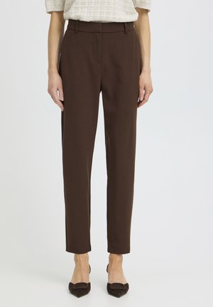 Frau trägt hoch taillierte, gerade geschnittene braune Hose, kombiniert mit braunen, spitz zulaufenden Schuhen und einem cremefarbenen Strickoberteil, steht vor weißem Hintergrund.