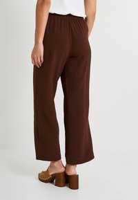 Une personne portant un pantalon large marron et des sandales à talons beiges est de dos, se tenant contre un fond blanc uni.