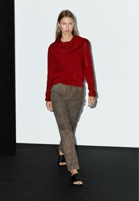 Vrouw die over de catwalk loopt in een rode gestructureerde coltrui, bruine suède broek, zwarte open teen hakken en een zilveren ring om haar rechterhand.
