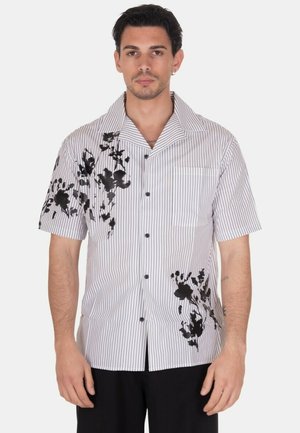Uomo che indossa una camicia bianca a maniche corte con sottili righe nere e motivi floreali neri sul lato sinistro e nella parte inferiore destra.