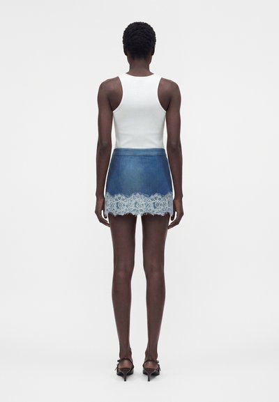 Άτομο στέκεται με την πλάτη, φοράει λευκό αμάνικο τοπ, μπλε τζιν μίνι φούστα με λευκή δαντέλα και μαύρα σανδάλια με τακούνι.