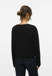 Pull noir en tricot avec une coupe décontractée, encolure ronde et manches longues, associé à un jean foncé. Le tissu semble texturé.