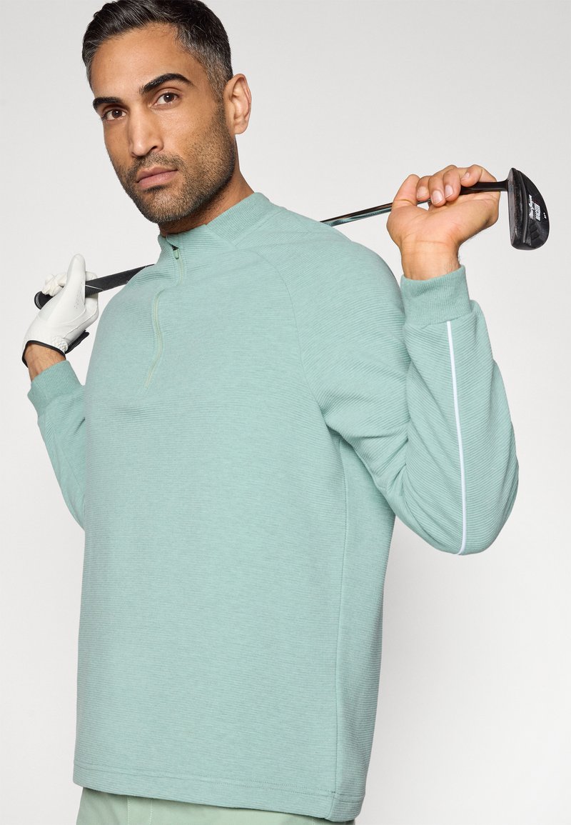 Pull-over vert clair à manches longues avec col à demi-fermeture éclair, présentant une texture côtelée et des bandes blanches en accent le long des manches.