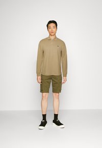 Khakifarbenes Poloshirt mit langen Ärmeln und Kragen, kombiniert mit olivgrünen Cargo-Shorts. Schwarze Sneakers mit weißen Sohlen. Sauberes, minimalistisches Design.