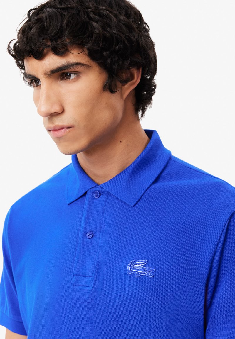 Royalblaue Poloshirt aus strukturiertem Stoff, mit klassischem Kragen und zwei Knöpfen, versehen mit einem gestickten Logo auf der linken Brust.