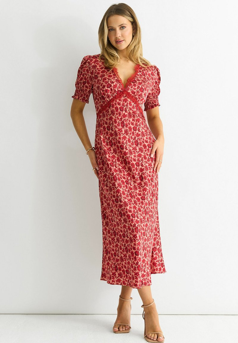 Red floral midi dress with short puff sleeves, delicate lace trim, and a fitted bodice, flowing to a loose hemline; paired with tan sandals.