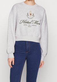 Pull court gris avec col rond, inscription brodée "Hôtel Mer" en vert, et poignets côtelés, associé à un jean bleu foncé.