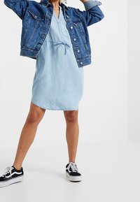 Donna che indossa un vestito azzurro chiaro, una giacca di jeans blu e sneakers in bianco e nero, in piedi con le gambe divaricate e le braccia sollevate.