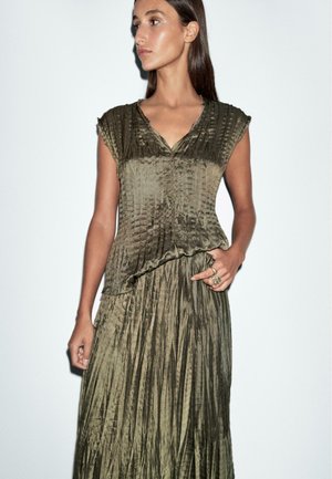 Robe vert olive plissée avec un col en V et des manches courtes. Fabriquée en tissu texturé. Le modèle porte des bagues à une main.