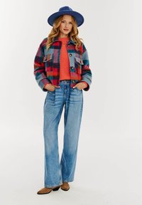 Farebný károvaný kabát z vlnenej zmesi s veľkými gombíkmi, skombinovaný s oranžovým sveterom a svetlo modrými širokými džínsami. Modrá čiapka dotvára celý outfit.
