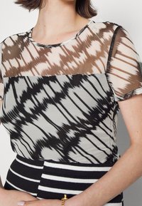 Maglietta a maniche corte con motivo tie-dye in tonalità di marrone, nero e crema. La parte superiore presenta dettagli in rete trasparente.