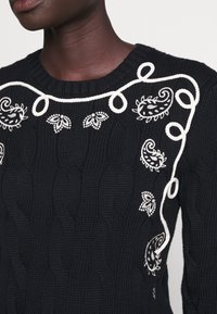 Pull en maille foncé avec des motifs paisley et floraux brodés blancs et un motif blanc en forme de corde près de l'encolure et de l'épaule.