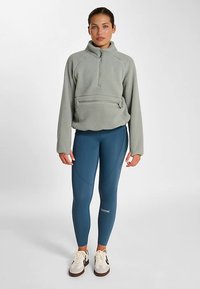 Pull polaire gris clair avec col à fermeture éclair et poche avant, associé à un legging bleu avec des coutures courbes et un détail logo.