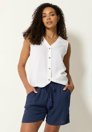 Femme aux cheveux bouclés portant un chemisier blanc sans manches boutonné et un short bleu marine à cordon avec des pompons, les mains dans les poches.