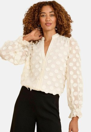 Blusa bianca con texture a pois, scollo a V, dettagli con bottoni e polsini arricciati. Le maniche sono lunghe e leggermente svasate. Indossata con pantaloni neri.