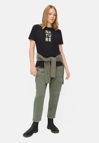Sort t-shirt med "NATURE" i fede, cremefarvede bogstaver; olivengrønne cargo-bukser; lys strikket sweater bundet rundt om livet; sorte ankelstøvler.