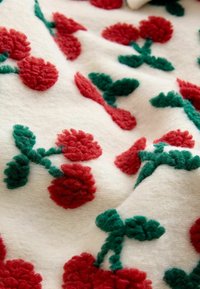 Bela tkanina z rdečim cvetličnim vezenjem in zelenimi listi. Tekstura je videti puhasta, z dvignjenim vezenjem, ki ustvarja tridimenzionalni učinek.