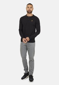 Zwarte poloshirt met lange mouwen en een ritskraag, voorzien van een klein gouden logo, gecombineerd met lichtgrijze broek en zwarte sportschoenen.