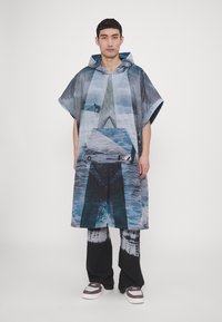 Hætteponcho med et foto af vand og geometriske mønstre i nuancer af blåt og gråt. Parret med tie-dye sorte bukser og sneakers.