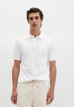 Junger Mann mit kurzen lockigen Haaren, der ein weißes kurzärmeliges Polohemd und beige Hosen mit Kordelzug trägt, steht vor einem schlichten hellen Hintergrund.