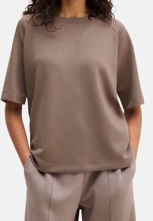 Vrouw draagt een losse taupekleurige top met korte mouwen en bijpassende taupekleurige broek met handen in de zakken tegen een effen achtergrond.