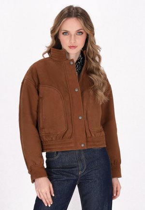 Donna con capelli lunghi e mossi indossa una giacca di suede marrone con bottoni a pressione e jeans blu scuro, in pose contro uno sfondo bianco.