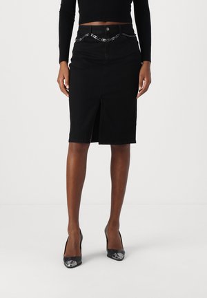 Femme portant une jupe noire longueur genoux avec une fente avant et une ceinture chaîne argentée, associée à des chaussures à talons noirs et un haut noir à manches longues.