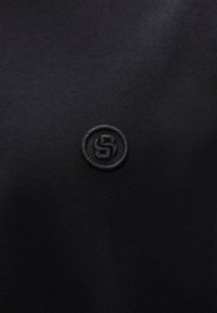 Fekete póló, amelyen a bal felső oldalon egy kicsi, kerek, fekete hímzett logó látható. Az anyag sima, minimális textúrával.