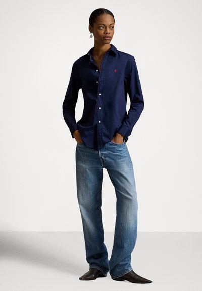 Ženska stoji z rokami v žepih, nosi mornarsko modro srajco z gumbi, svetlo modre jeans hlače in črne koničaste čevlje na preprostem belem ozadju.