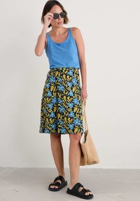 Top bleu fără mâneci, asociat cu o fustă până la genunchi care are un model floral colorat în nuanțe de albastru și galben pe un fundal negru. Sandale negre și o geantă de umăr bej.